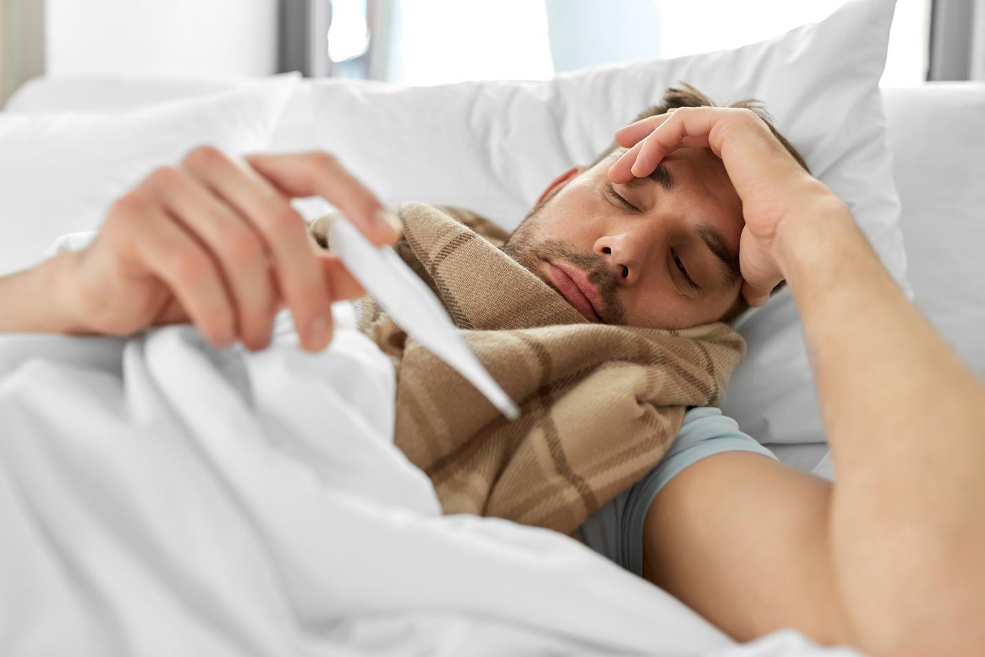Ein Mann liegt im Bett, schaut auf ein Thermometer, ist in einen Schal gehüllt und hält sich den Kopf