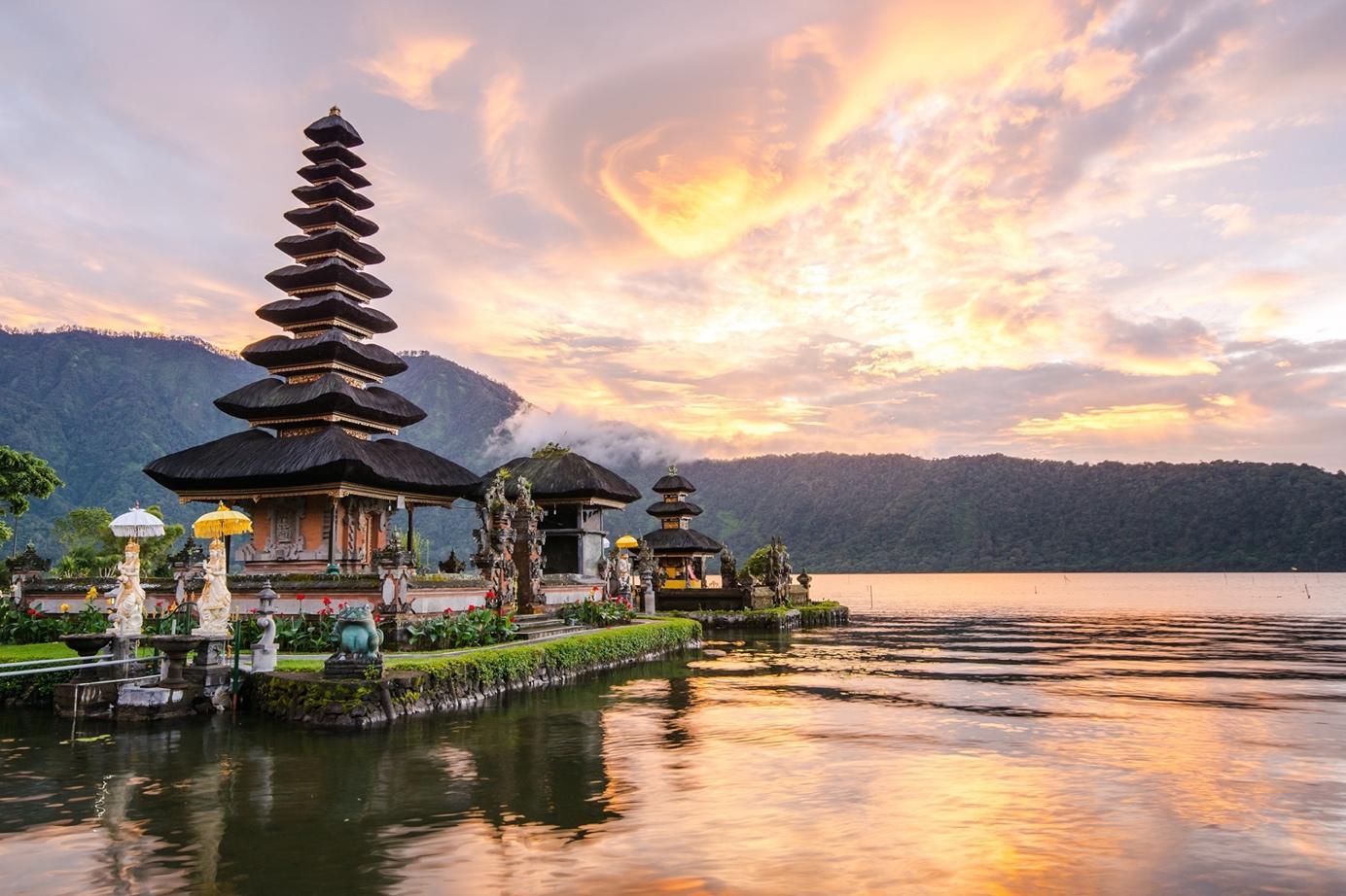 Ein Tempel in Bali sitzt über dem Wasser, während die Sonne hinter hohen Bergen in der Ferne untergeht