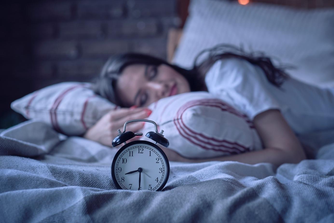 Eine junge Frau schläft im Bett und hält ihr Kissen und ein Wecker sitzt auf dem Bett vor ihr