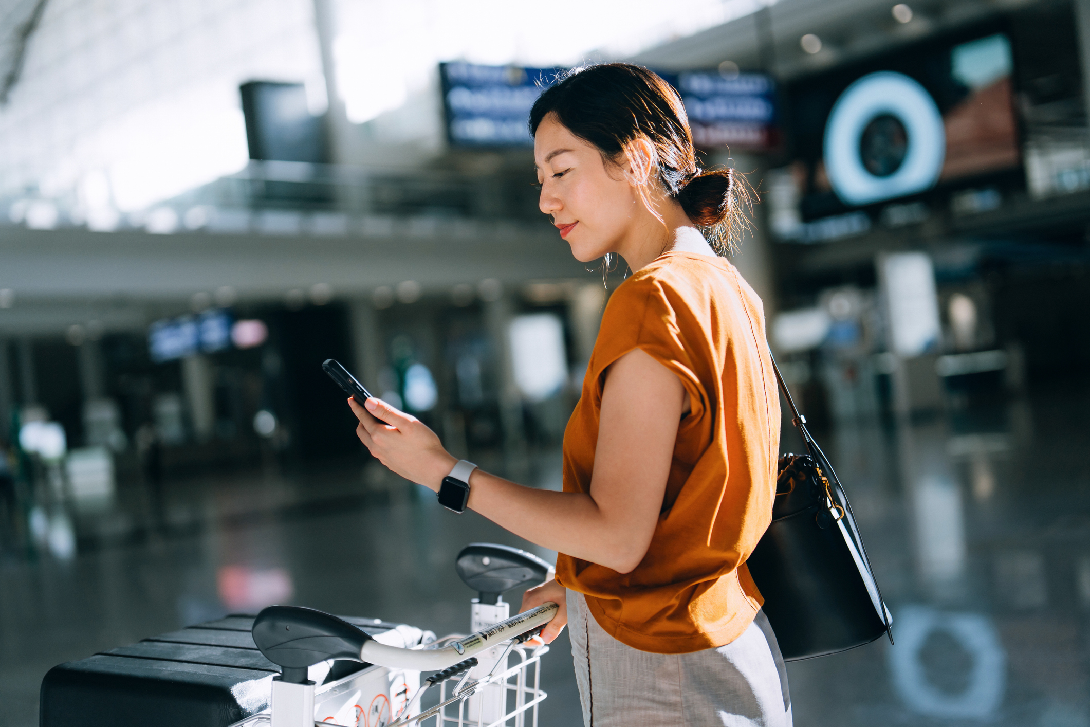 Une femme tient un smartphone dans sa main gauche et le regarde en souriant. Elle est entourée d'un paysage urbain. Un ruban violet est visible à l'arrière-plan de l'image.