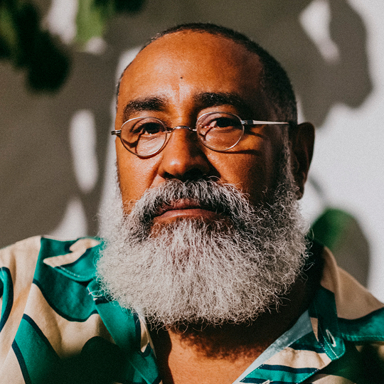 Portrait d'un homme à la peau brune, portant de petites lunettes à monture et une barbe grise. Il porte une chemise à motifs verts et crème.