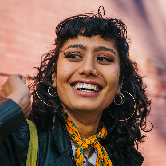 Photo d'une femme souriante, avec des cheveux courts, foncés et bouclés. Elle porte un foulard jaune autour du cou et une veste bleu foncé.