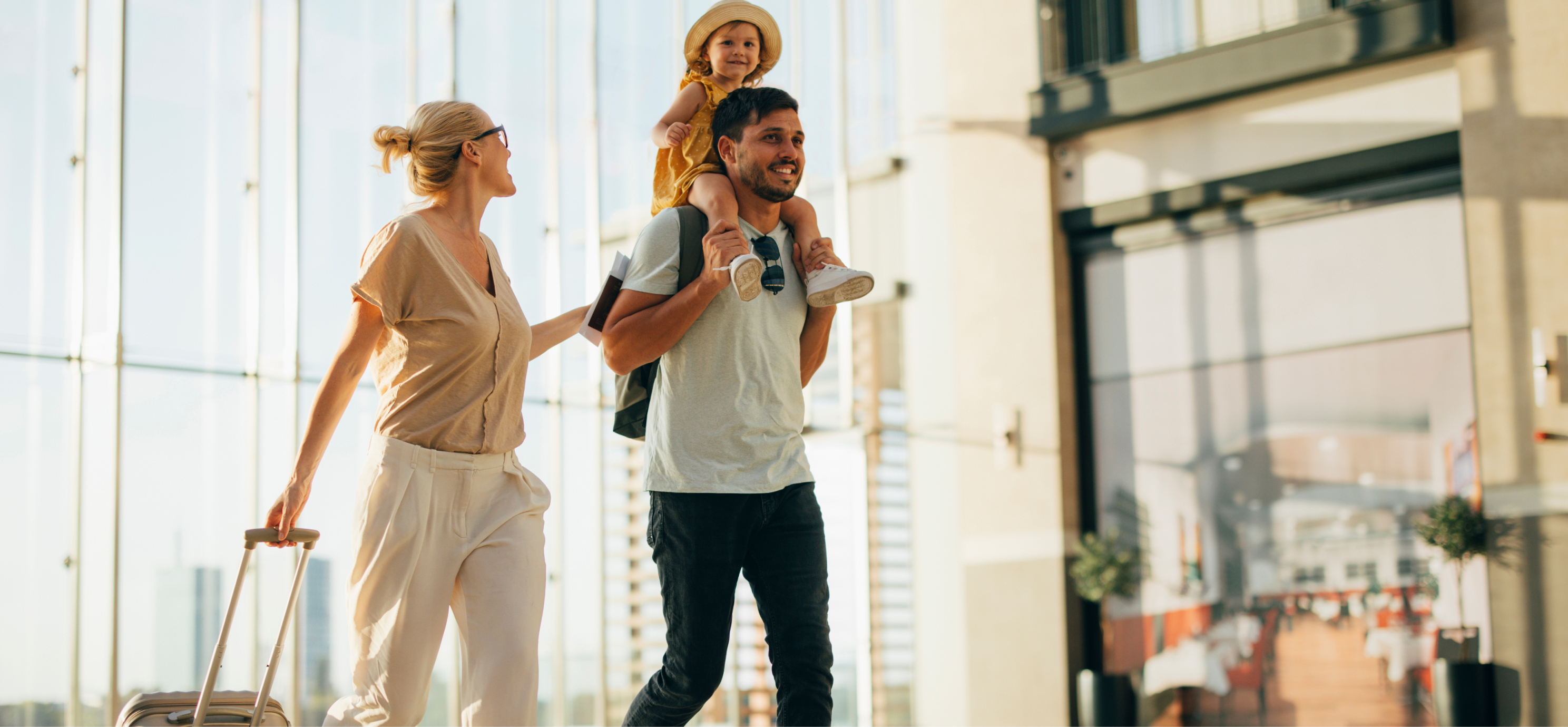 Une femme à la peau claire, aux cheveux blonds et portant des lunettes tire une valise. Elle a une main posée sur son partenaire, un homme à la peau brun clair et à la barbe. Un enfant portant un chapeau de soleil est assis sur ses épaules.